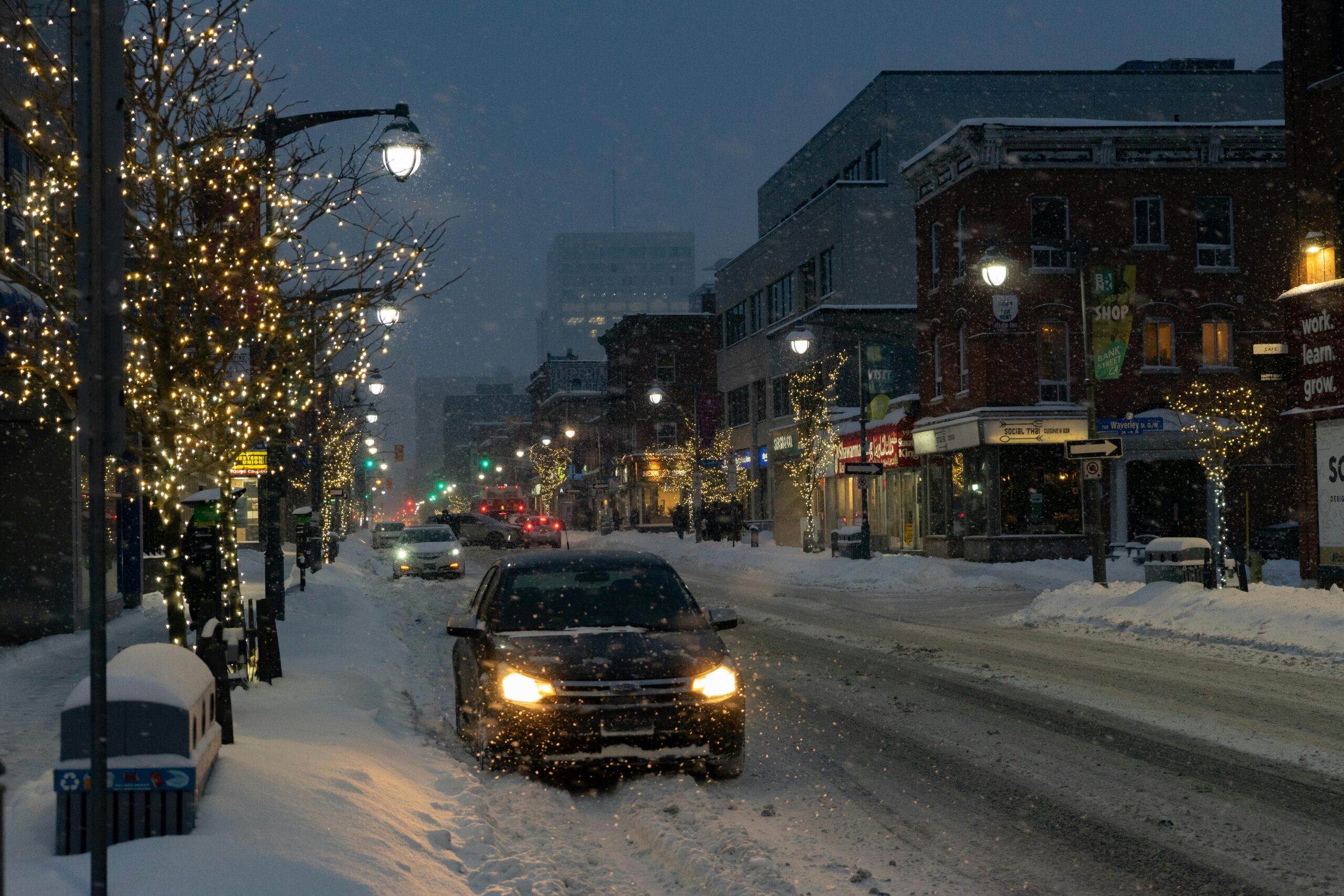 Snowy street scene during holiday season with cars navigating through winter weather, festive lights illuminating storefronts, and potential driving hazards.