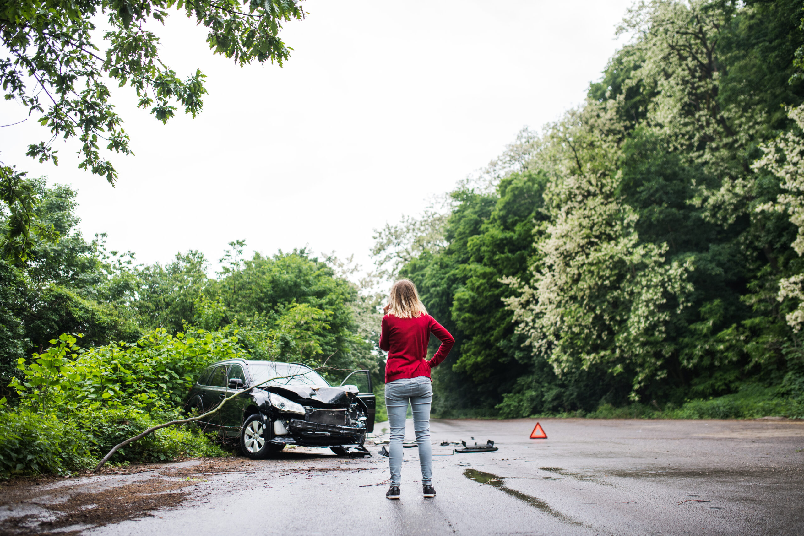Young woman in red sweater standing by a damaged car on a road, surrounded by greenery, highlighting the dangers of driving on wet leaves.