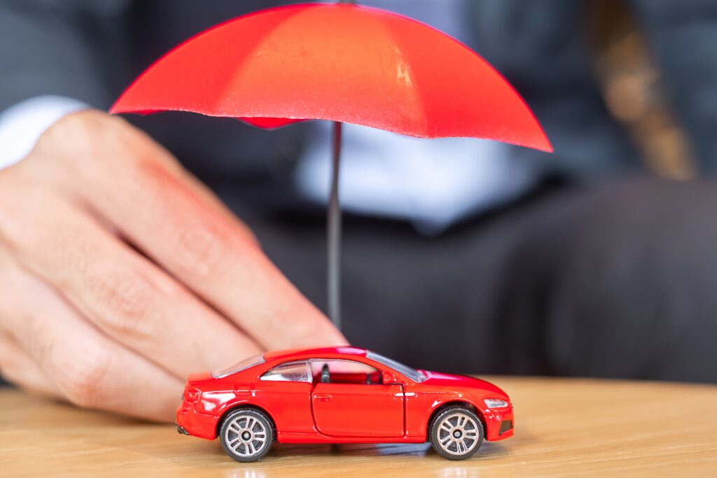 Businessman&rsquo;s hand holding a red umbrella over a toy car, symbolizing insurance protection and coverage.