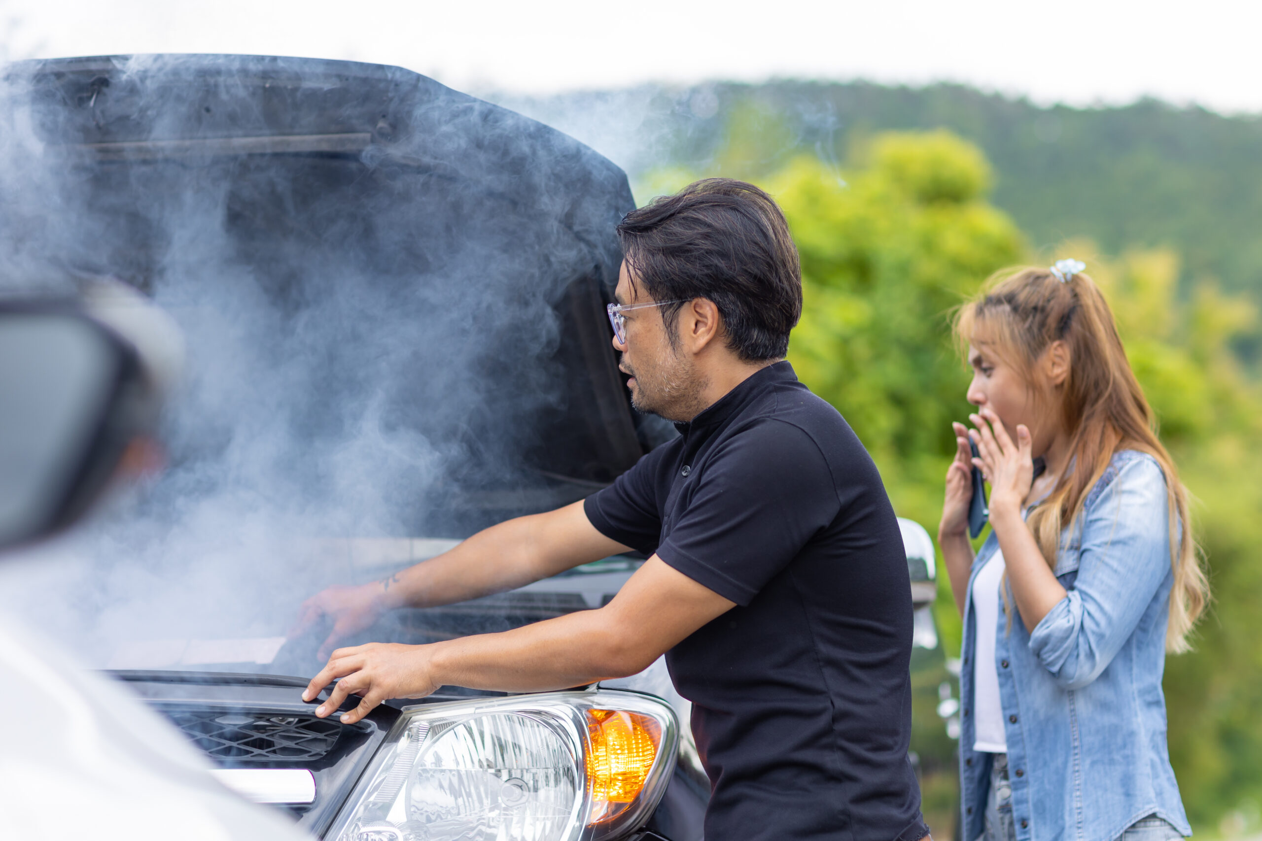 Man inspecting a smoking car engine with open hood, woman reacting in concern, summer driving context highlighting engine overheating risks.