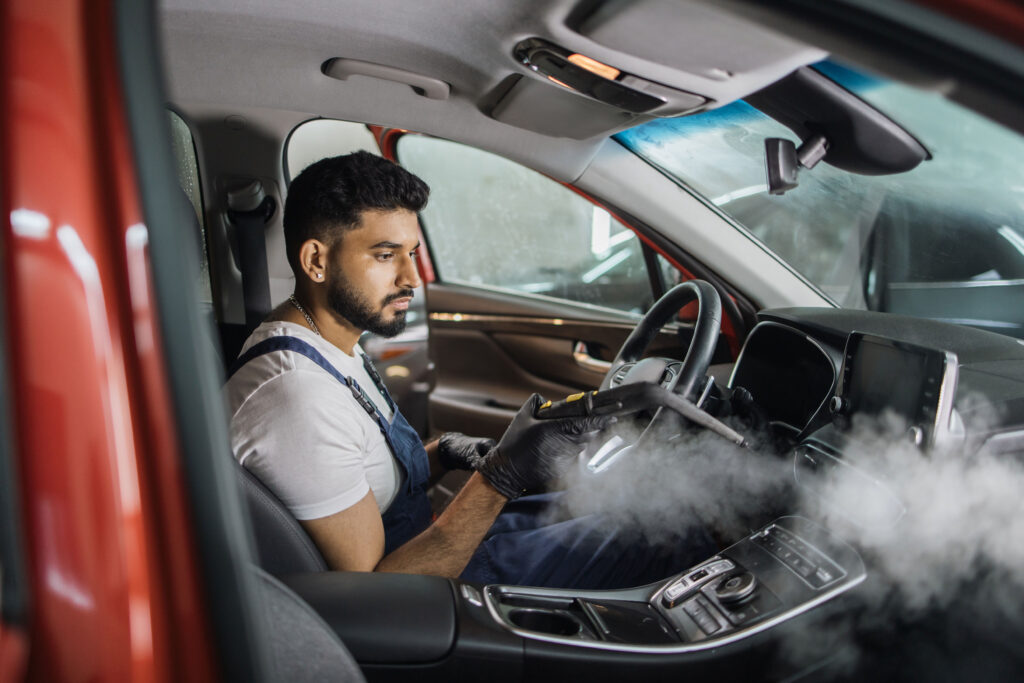 Man in blue overalls cleaning car interior with steam cleaner, emphasizing car maintenance and odor removal after road trips.