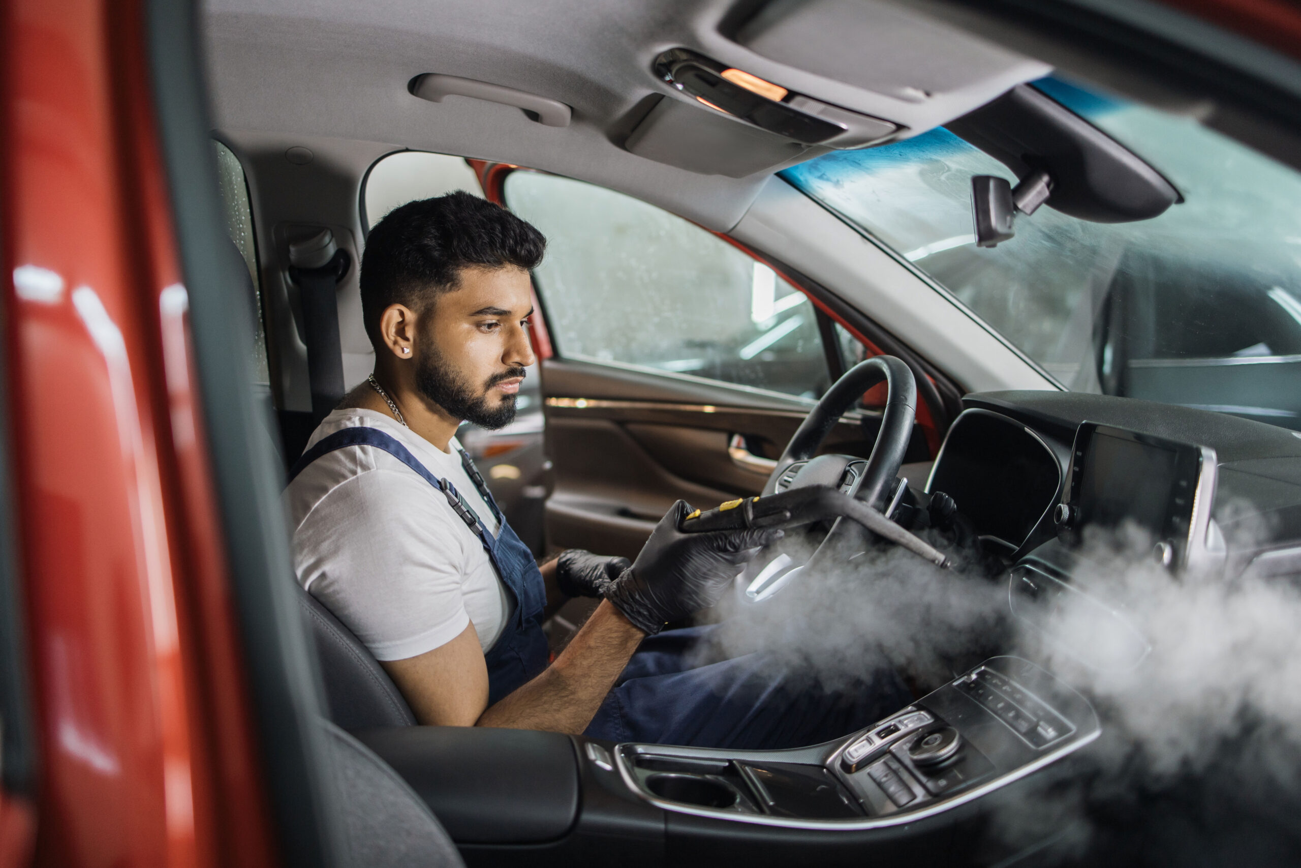 Man in blue overalls using a steam cleaner inside a car, illustrating maintenance and cleaning related to automotive odors.