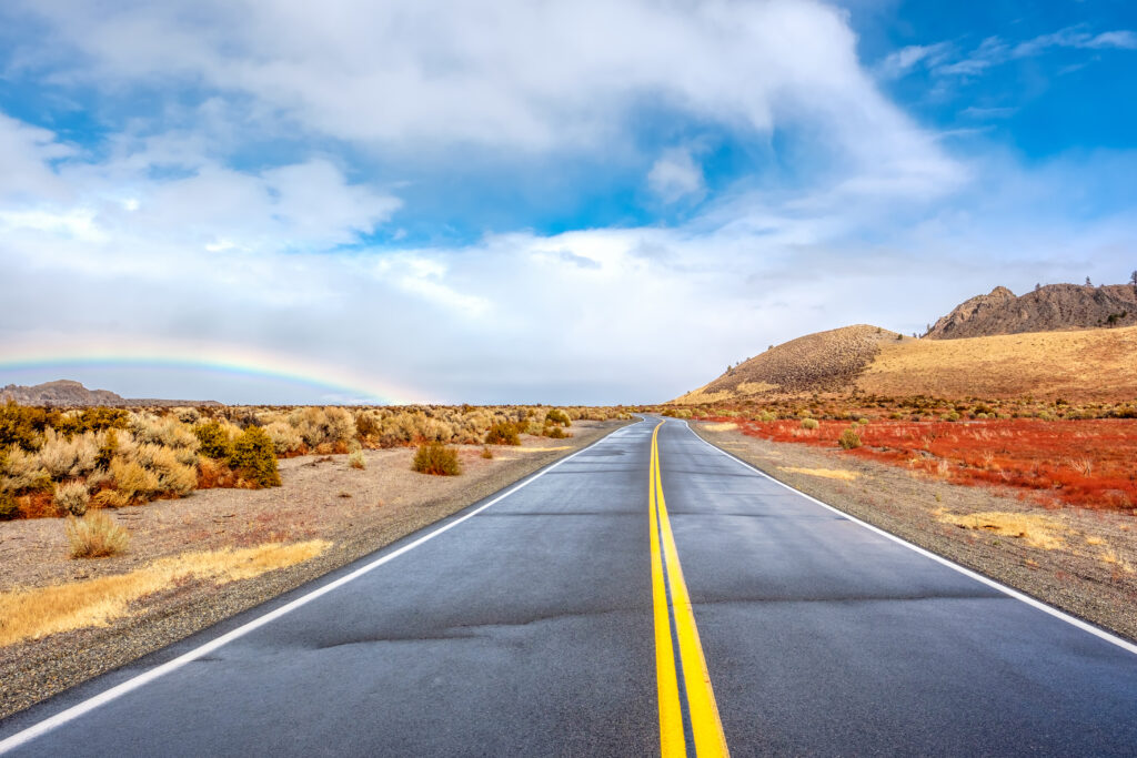 Open highway with a rainbow in the sky, surrounded by dry landscape and mountains, symbolizing freedom and the journey of safe driving after long trips.