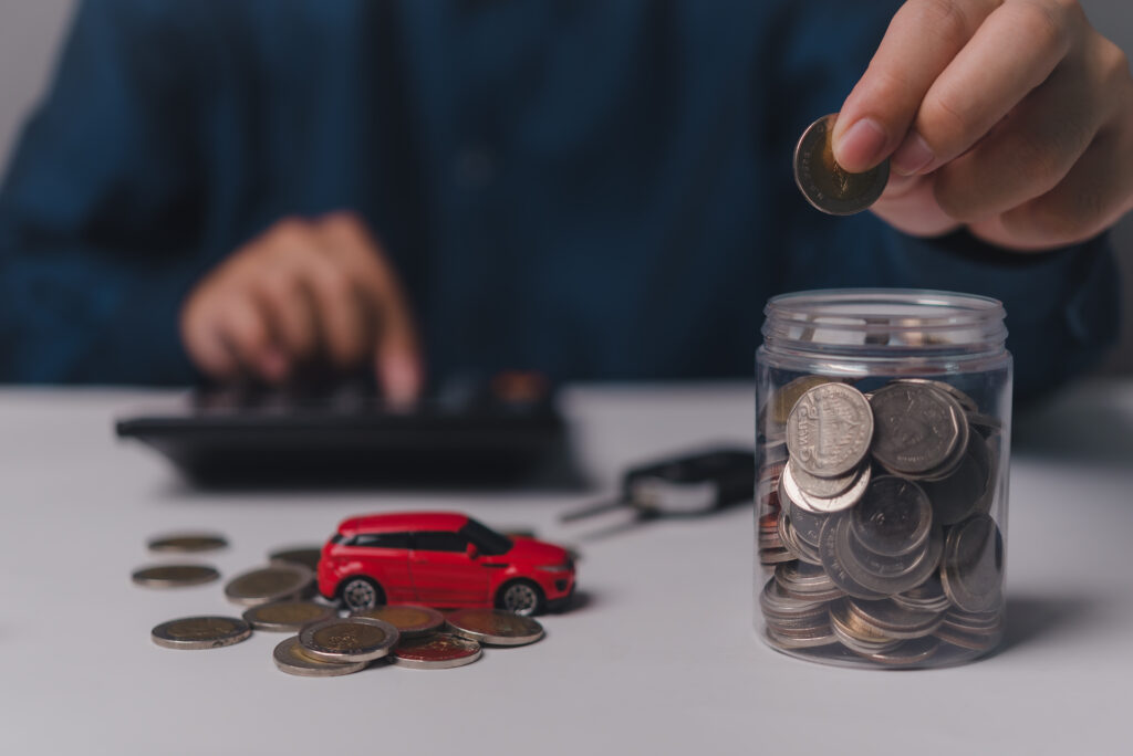 Person placing coin into jar while calculating savings for car insurance, with toy car and coins on table, illustrating financial planning for insurance review.