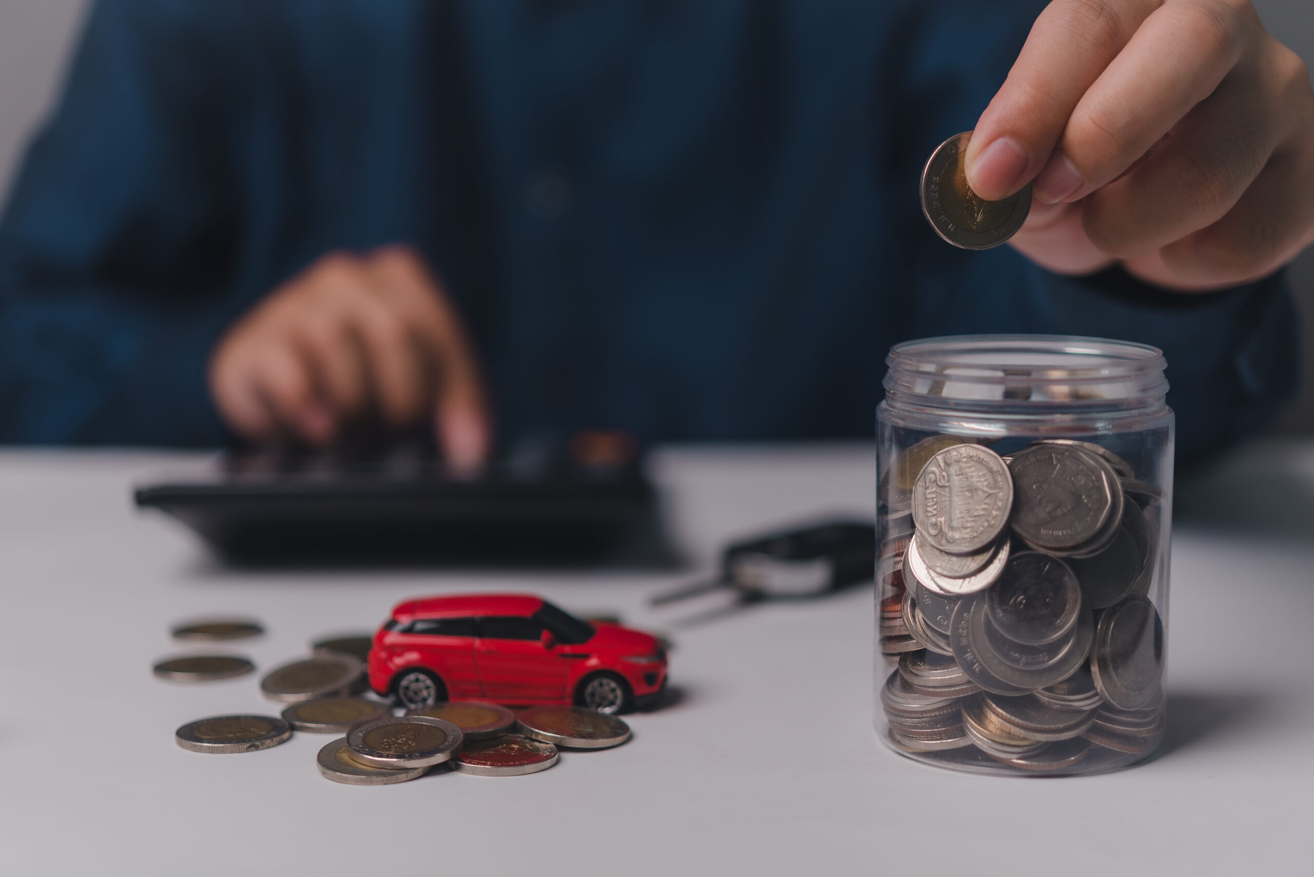 Person adding coins to a jar while a toy car and scattered coins are on the table, illustrating savings for car expenses related to insurance review.