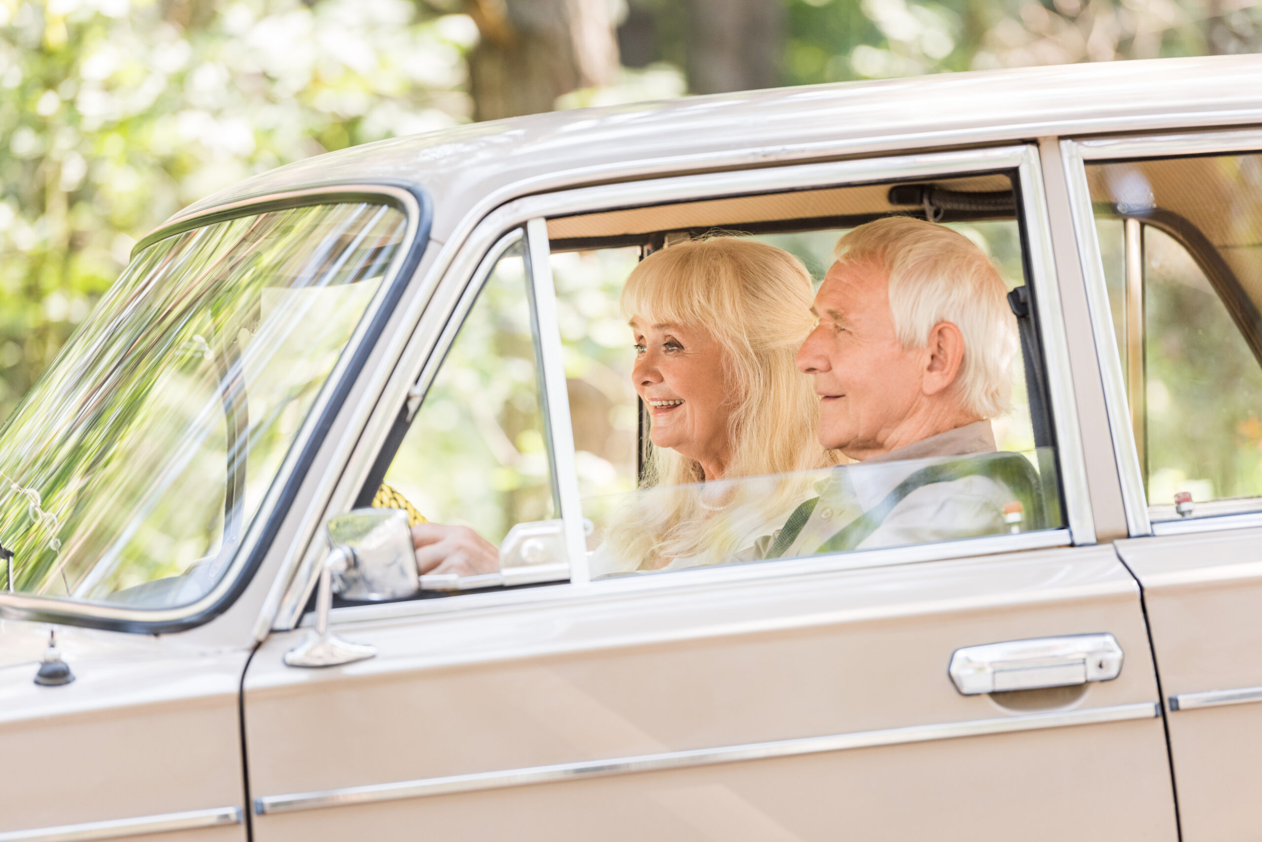 Senior couple driving in a beige car, smiling and enjoying the ride, surrounded by a natural setting, emphasizing safe driving for older adults during seasonal changes.