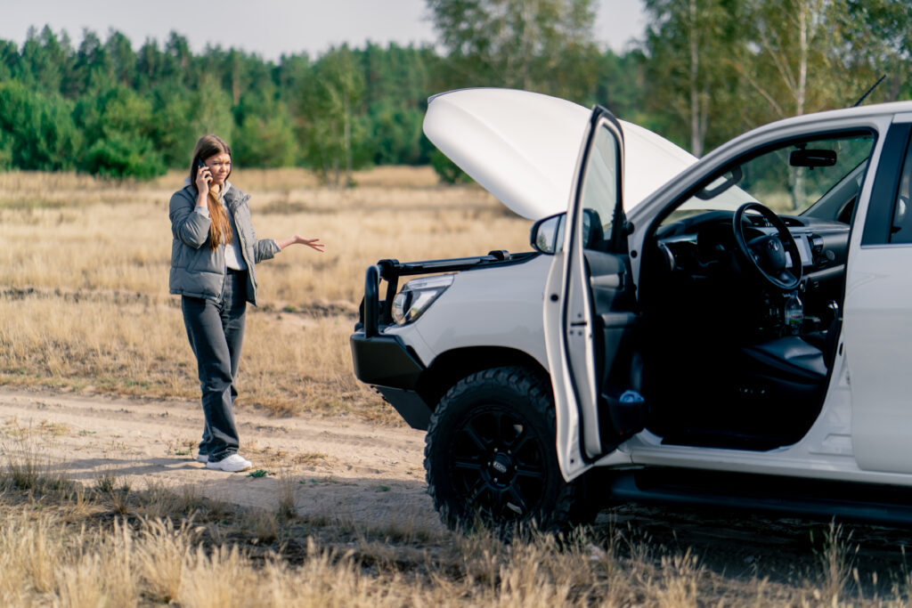 Young woman on phone beside broken-down car in forest, emphasizing road trip challenges and potential pitfalls.