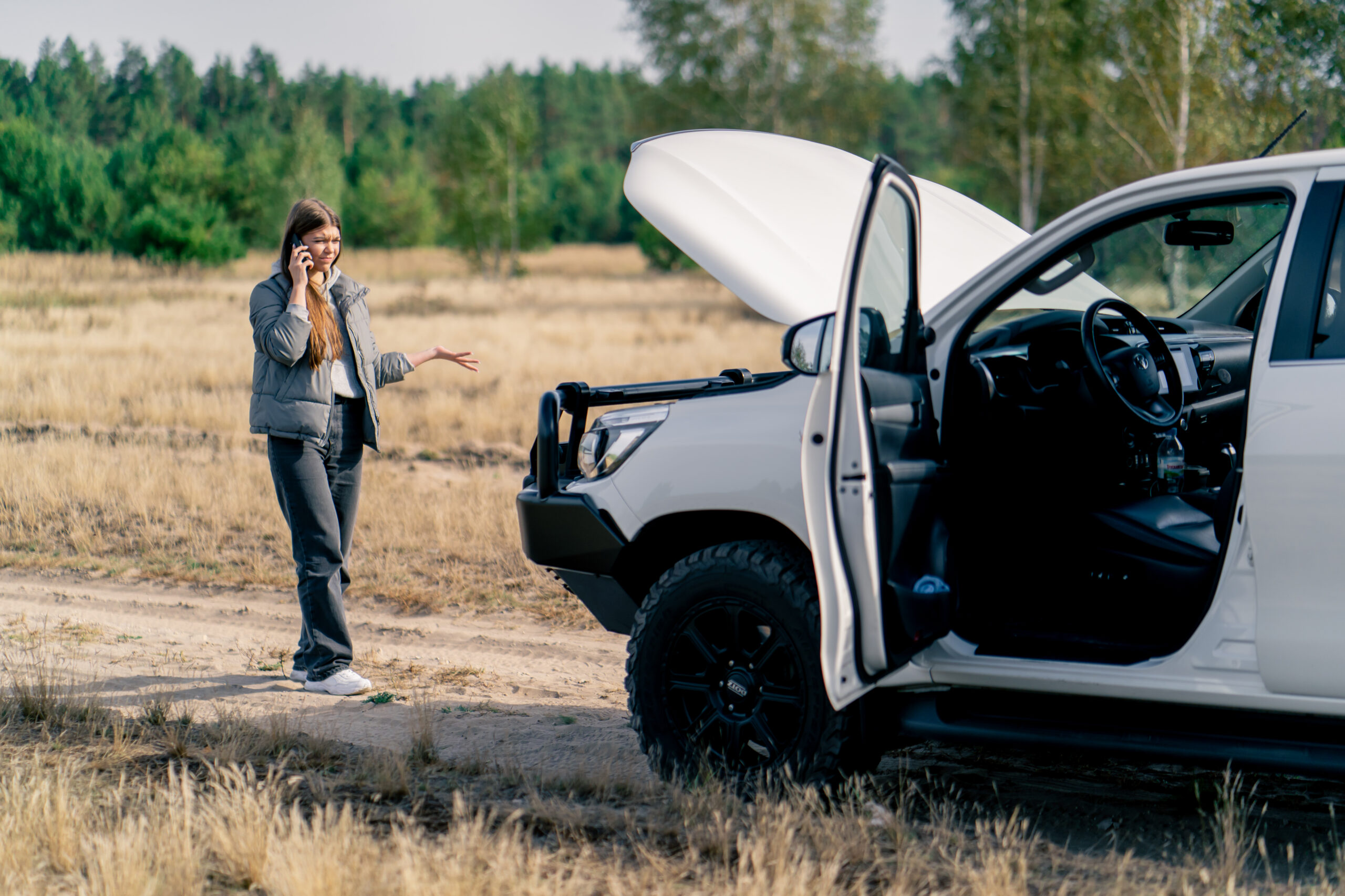Young woman on phone near broken-down car with hood raised in forested area, illustrating road trip mechanical issues and the importance of preparation for safe travel.