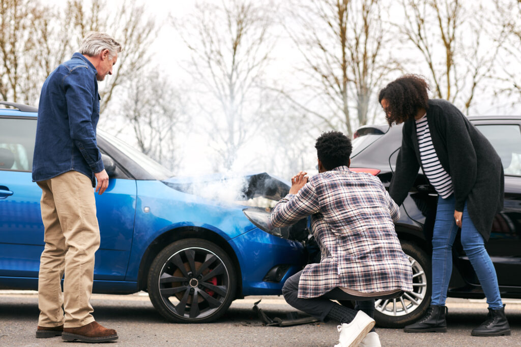 Young couple inspecting a blue car with steam rising from the hood, indicating engine overheating, while an older man observes nearby, reflecting a roadside breakdown situation.