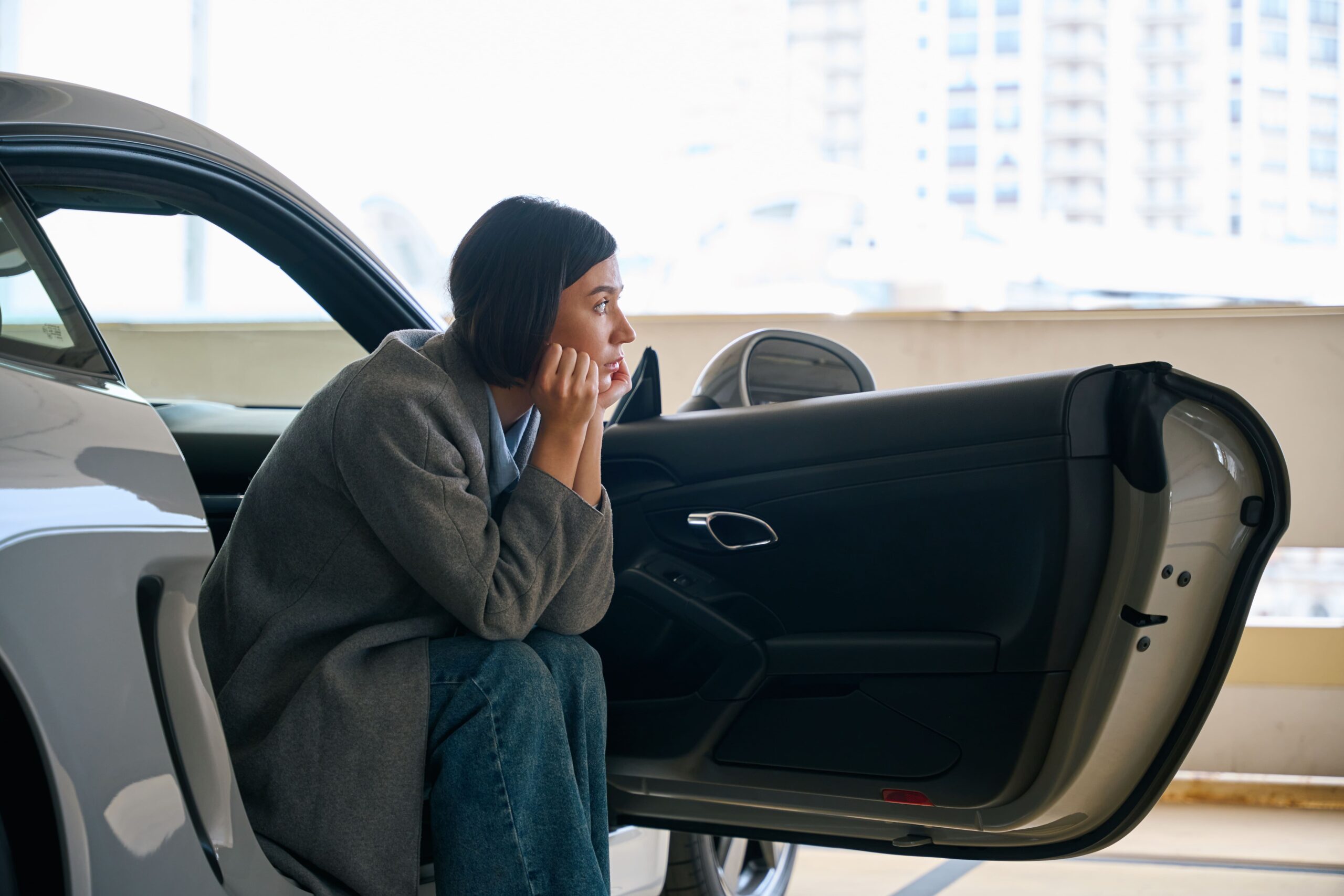 Woman sitting in her car seat with the door open, her head resting on her hands, gazing thoughtfully into the distance